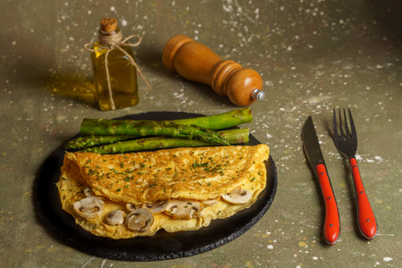 omelette stuffed with mushrooms and asparagus on a black slate plate with olive oil, pepper and cutlery.の写真素材