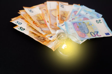 illuminated light bulb with euro banknotes in the background on a black background, concept of rising electricity pricesの写真素材
