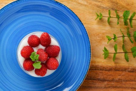 raspberries with yogurt in a glass bowl and a blue plate on a wooden table