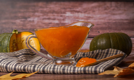 homemade pumpkin jam in a glass jar on a wooden tray with dried leaves with cut pumpkin on the bottomの写真素材