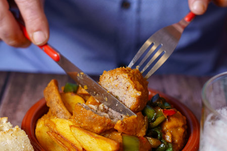 man in blue shirt cutting a meatball with fork and knife into a portion in an earthenware dishの写真素材