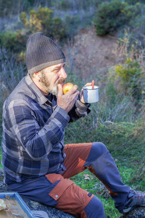 man with beard and black cap drinking coffee and eating an apple in the fieldの写真素材