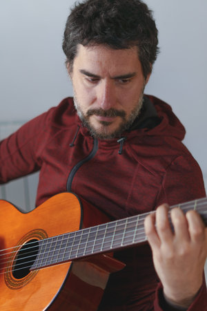 young man with beard playing Spanish guitar looking at the strings of his guitar with white...