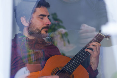 young man with beard playing guitar seen through windowの写真素材