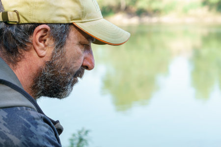 bearded fisherman with cap and beard seen in profile on the river bank carp fishing sessionの写真素材