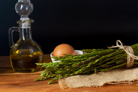 bunch of wild asparagus on a raffia cloth with a jar of olive oil and eggs black backgroundの写真素材
