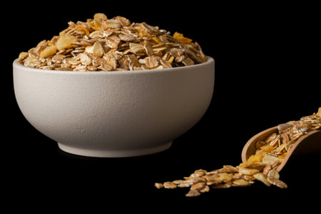 close-up of a white bowl of muesli with wooden spoon with flakes isolated on black background and...