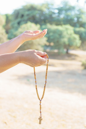 woman's hands holding a Christian rosary with a cross, praying in a field at duskの写真素材