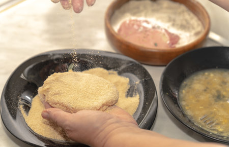 A woman's hand dips a pork fillet into breadcrumbs. In the background you can see the flour and egg...