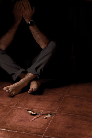 unrecognizable barefoot man in semi-darkness sitting on the floor with a syringe and spoon with heroin in the foregroundの写真素材