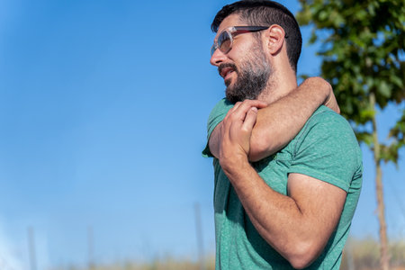 profile view of an attractive bearded man with sunglasses stretching his shoulders in a field with blue sky in the backgroundの写真素材