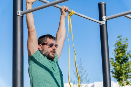 Bearded man with sunglasses seen in profile training his back by doing pull-ups on a barbell with an...