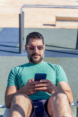 young man with a beard and sunglasses consults an exercise application on his mobile phone while working out in a gym in the park.の写真素材