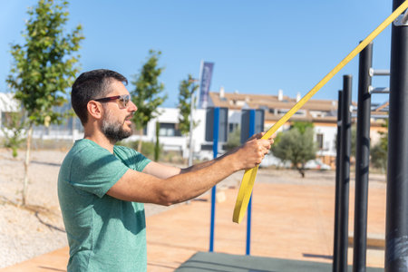 Bearded man with sunglasses seen in profile training his arms with an elastic fitness band in an...