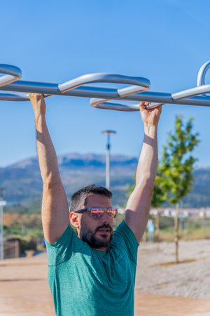Bearded man with sunglasses seen from the front training his back by doing pull-ups on a barbell assisted by an elastic fitness band in an outdoor gym.の写真素材