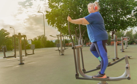 active obese elderly woman in sportswear working out on a park machine while listening to music on her headphonesの写真素材