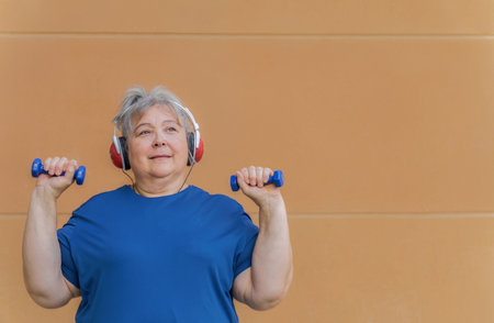 White haired obese older woman exercising with dumbbells and headphones listening to music for weight loss .Isolated on orange background and copy space.の写真素材