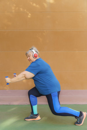 White haired obese older woman exercising with dumbbells and headphones listening to music for weight loss .Isolated on orange background and copy space.の写真素材