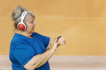 Older woman with overweight, sportswear and headphones consults her smartwatch while training in the park.の写真素材