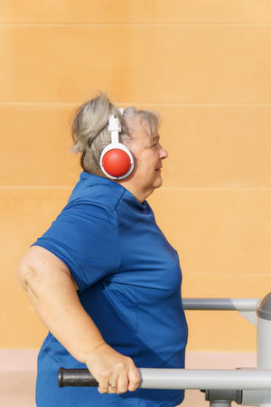 Profile view of an active obese elderly woman in sportswear working out on a gym machine in a park gym while listening to music on her headphonesの写真素材