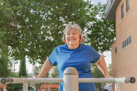 active older woman in sportswear working out on a gym machine in the park while listening to music...