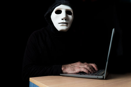 hacker dressed in black with mask and hood typing on a computer while looking at the camera cybercrime conceptの写真素材