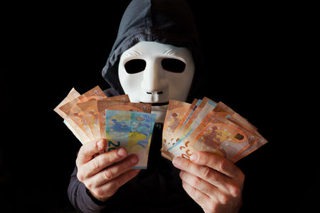 hacker man dressed in black with white mask and black hood counting euro banknotes in his hands, cybercrime concept, isolated on black backgroundの写真素材