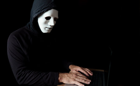 male hacker dressed in black with a hood and a white mask using a computerの写真素材