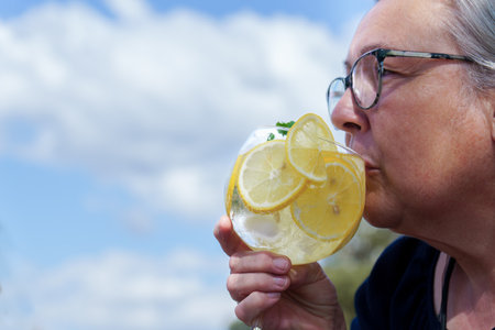 woman seen in profile drinking a glass of gin and tonic with lemon slices and mint leaves with a blue sky with clouds in the backgroundの写真素材