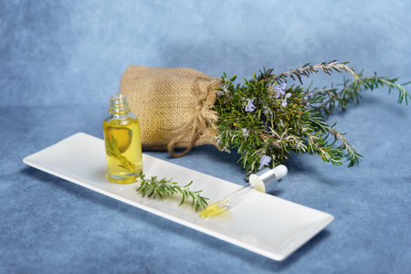 dropper with rosemary essential oil with fresh branches inside on a white tray next to a burlap bag with flowering rosemary branchesの写真素材