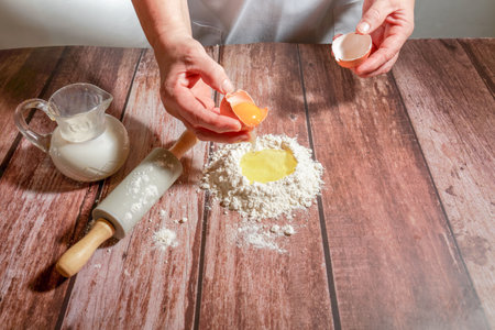 woman breaking an egg, pouring it over flour to make a sponge cake on a wooden table with a rolling pin and a pitcher of milkの写真素材