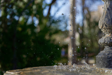 profile view of a public fountain with splashing water, blue sky and park trees in the background.の写真素材