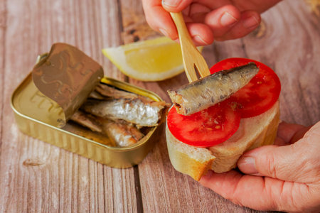 a woman puts a sardine on a slice of bread with tomato, in the background you can see the can of sardines and a lemon on a wooden table.の写真素材