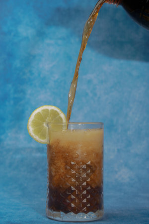 a cola is poured into a glass on a glass with ice and lemon on a blue background with copy spaceの写真素材