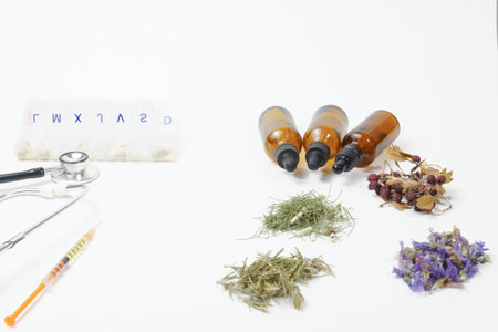 In the foreground, a stethoscope, a syringe and a box of pills rest next to bottles of essential oils and bunches of medicinal herbs on a white table, naturopathy concept.の写真素材