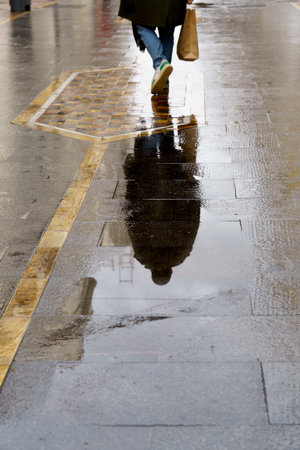 woman with shopping bags walking down a street reflected in a puddle on a rainy day