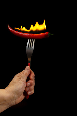 close-up of woman's hand holding a fork with a hot chili burning in flames, isolated on black background and copy spaceの写真素材