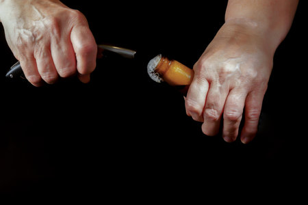 Close-up of wrinkled hands opening a beer bottle with a knife, set against a black backgroundの写真素材