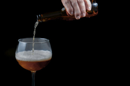 Hand pouring beer from a bottle into a glass, creating foam, on a black backgroundの写真素材