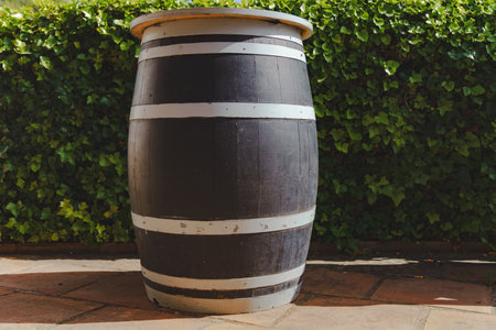 Large, dark-colored wine barrel with white stripes stands on a tiled floor near a green hedge, creating a rustic atmosphereの写真素材