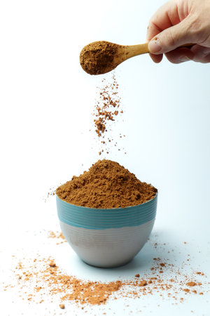 Cocoa powder falling from wooden spoon into bowl on white background, creating a cloud of dust