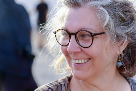 Close-up of cheerful mature woman with eyeglasses, enjoying golden hour sunlight, showing a genuine...