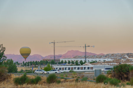 Hot air balloon flying over a construction site with cranes and houses at sunsetの写真素材