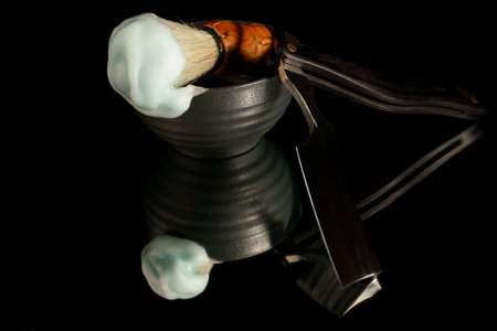 Classic straight razor, bowl, and brush with foam reflecting on a dark surfaceの写真素材