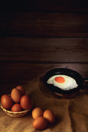 Rustic still life featuring a single fried egg in a small pan flanked by fresh brown eggsの写真素材