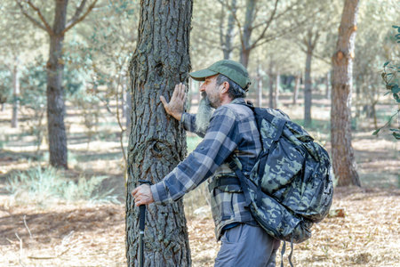 Man in plaid shirt and trekking backpack touching the bark of a tree in a forest, finding connection...