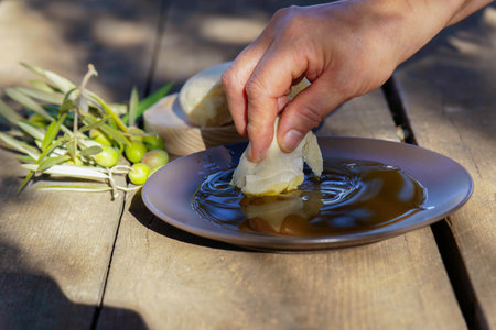 Hand dipping a piece of fresh bread into a shallow plate of extra virgin olive oil, with olives and olive branches on a rustic wooden tableの写真素材