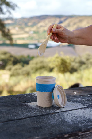 Person's hand adding a white powder with a wooden spoon into a reusable eco-friendly cup, enjoying a...