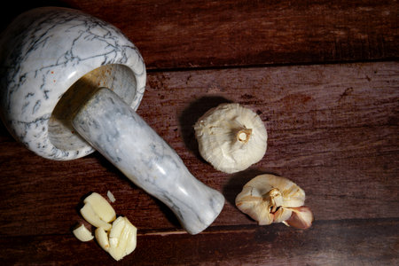 Marble mortar and pestle lying next to whole garlic bulbs and peeled cloves on a rustic wooden table, preparing ingredientsの写真素材