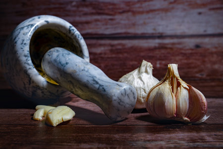 Food preparation scene featuring fresh garlic bulbs and peeled cloves next to a marble mortar and pestle on a dark wood surfaceの写真素材